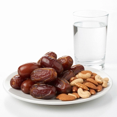 A variety of fresh dates arranged aesthetically on a white plate, with some nuts and a glass of water in the background, suggesting a healthy snack.
